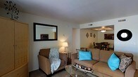 This single-floor residence offers a welcoming and well-appointed layout designed for comfortable everyday living. The living area serves as the heart of the home, featuring plush seating including a cushioned armchair draped with a decorative throw and a spacious sectional sofa accented with vibrant teal pillows. A glass-topped coffee table anchors the seating arrangement, while a stylish floor lamp provides warm ambient lighting. A large framed mirror on the wall adds depth and brightness to the room, complementing the clean, neutral-toned walls. Decorative diamond-shaped wall accents create a subtle artistic touch that carries through to the adjoining dining area. The open-concept flow connects the living space seamlessly to the dining area, which is furnished with a classic dining table and chairs beneath a pendant light fixture, making it ideal for both casual meals and entertaining. Carpeted flooring runs throughout the living space, adding warmth and comfort underfoot. Ceiling vents ensure year-round climate control. The overall ambiance is cozy and inviting, with a cohesive neutral palette punctuated by tasteful decorative accents that give the space a lived-in yet polished feel.