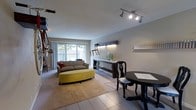 This single-floor space combines a living area and dining nook in an open, functional layout. The living area is anchored by a grey sofa paired with a chartreuse oval ottoman atop a neutral area rug, creating a relaxed and inviting atmosphere. Large sliding patio doors along one wall flood the space with natural light and offer views of an outdoor patio and greenery beyond, enhancing the sense of openness.

Track lighting mounted on the ceiling provides targeted illumination throughout the space. The flooring transitions from large-format light tiles near the dining area to wall-to-wall carpet in the living zone. A sleek dark console table with under-shelf storage serves as a focal point along one wall, decorated with a ceramic vase and small greenery accents, with subtle LED strip lighting beneath.

The dining area features a compact dark wood table with ornate Queen Anne-style chairs, suited for intimate gatherings. A wall-mounted spice rack and labeled shelving nearby suggest proximity to a kitchen. The overall ambiance is casual yet thoughtfully styled, with neutral tones and pops of color creating a welcoming, livable environment.