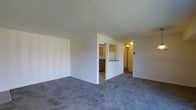 This single-floor residence offers a straightforward and functional layout designed to maximize everyday comfort. The living area is finished with wall-to-wall gray carpeting that adds a soft, neutral foundation to the space, while clean white walls provide a bright and airy atmosphere throughout. A half-wall partition elegantly separates the living area from the kitchen, creating a sense of openness while maintaining a defined layout. The kitchen features warm wood-toned cabinetry, light countertops, and tile flooring, giving it a classic and practical character. A pass-through counter connecting the kitchen to the living space enhances the open feel and allows for easy interaction between the two areas. A hallway extends beyond the kitchen, leading to additional rooms and providing a logical flow through the home. Overhead lighting in the kitchen and hallway ensures each area is well-lit. The overall design is clean and understated, offering a versatile canvas that can be easily personalized to suit a variety of interior styles. The single-floor plan makes the entire space easily accessible and convenient to navigate.