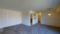 This single-floor residence offers a straightforward and functional layout designed to maximize everyday comfort. The living area is finished with wall-to-wall gray carpeting that adds a soft, neutral foundation to the space, while clean white walls provide a bright and airy atmosphere throughout. A half-wall partition elegantly separates the living area from the kitchen, creating a sense of openness while maintaining a defined layout. The kitchen features warm wood-toned cabinetry, light countertops, and tile flooring, giving it a classic and practical character. A pass-through counter connecting the kitchen to the living space enhances the open feel and allows for easy interaction between the two areas. A hallway extends beyond the kitchen, leading to additional rooms and providing a logical flow through the home. Overhead lighting in the kitchen and hallway ensures each area is well-lit. The overall design is clean and understated, offering a versatile canvas that can be easily personalized to suit a variety of interior styles. The single-floor plan makes the entire space easily accessible and convenient to navigate.