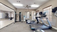 This fitness center is designed to support a comprehensive workout experience within a single-floor layout. The space is finished with neutral taupe walls, low-pile carpet flooring, and white trim, creating a clean and functional atmosphere throughout. Overhead fluorescent lighting, supplemented by ceiling fans, ensures the room remains well-lit and comfortable during use.

The cardio area is well-appointed with multiple treadmills, an elliptical trainer, and a stair-climber, each equipped with individual display screens for tracking workout metrics. A wall-mounted television provides additional entertainment options for users. A full cable weight machine with suspension training attachments occupies one side of the room, offering a range of strength training exercises, while a dumbbell rack lines the mirrored wall, which visually expands the space and aids in form monitoring.

A floor-to-ceiling glass partition separates an adjacent room, adding visual openness. Multiple windows fitted with wood-toned blinds allow natural light to filter in while maintaining privacy. A water cooler station is conveniently positioned within the space. Posted workout guides and informational signage further enhance the usability of this well-organized fitness facility.