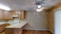 This single-floor space presents a straightforward, functional layout with a cohesive design throughout. Neutral, warm-toned walls create a consistent aesthetic across the rooms, complemented by hardwood flooring that extends through the main living and kitchen areas, while carpet appears in at least one adjoining room.

The living area connects openly to an adjacent room through a wide doorway, offering a sense of flow between spaces. Ceiling fans with integrated light fixtures are installed in multiple rooms, providing both ventilation and general lighting. Textured popcorn ceilings are a consistent architectural detail throughout the unit.

The kitchen features medium-toned wood cabinetry, a white electric range with an overhead ventilation hood, and light-colored countertops. The kitchen opens toward the dining or living area, making it practical for everyday use. White baseboards trim the rooms throughout, adding a clean contrast to the warm wall tones.

Natural light enters through windows and a vertical blind-covered door visible from the main room. Overall, the space maintains a simple, utilitarian character with consistent finishes and a connected, easy-to-navigate single-level floor plan.