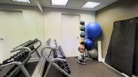 This single-floor fitness space is purpose-built for effective strength and functional training. The room features a drop ceiling with recessed fluorescent lighting that provides bright, even illumination throughout. A full-length wall mirror on one side enhances the sense of space while allowing users to monitor their form during exercise. The flooring is a durable, dark rubber-style gym carpet suited for heavy equipment and high-traffic use. A multi-tier dumbbell rack holds a comprehensive range of free weights, catering to various fitness levels and exercise types. Adjacent to the weights, a vertical medicine ball rack displays an assortment of weighted balls in varying sizes, ideal for core training and dynamic movements. A collection of exercise and stability balls, including a large blue stability ball, are stored on a dedicated ball rack alongside a foam roller, supporting stretching and balance work. The neutral, light-colored walls keep the environment clean and distraction-free, while the efficient use of floor space ensures that multiple pieces of equipment are accessible within a compact footprint. Overall, the room offers a focused and functional training environment well-suited for individual or small-group workouts.