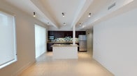 This single-floor residence offers a thoughtfully designed open-concept layout that seamlessly connects the kitchen and living areas, creating a spacious and airy atmosphere. The kitchen is a standout feature, showcasing rich dark wood cabinetry that contrasts beautifully against a light-toned tile backsplash with subtle mosaic accents. Stainless steel appliances, including a refrigerator and built-in microwave, add a sleek, contemporary feel. A central kitchen island with a stone countertop provides additional prep space and serves as a natural gathering point. Track lighting mounted on the ceiling illuminates the space efficiently, while exposed beam detailing adds architectural character to the white ceiling. Light-toned large-format floor tiles run throughout, contributing to a clean and cohesive aesthetic. A large frosted window allows diffused natural light to fill the space while maintaining privacy. A structural column near the island integrates naturally into the open floor plan, and a hallway visible from the kitchen suggests additional rooms beyond, completing this well-appointed and functional single-level home.