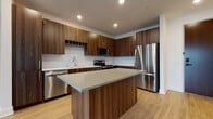 This single-floor residence showcases a contemporary design with a cohesive palette of warm wood tones, light hardwood flooring, and crisp white finishes throughout. The kitchen serves as a standout feature, equipped with rich walnut-grain cabinetry extending floor to ceiling, a herringbone-patterned tile backsplash, and a full suite of stainless steel appliances including a refrigerator, range, microwave, and dishwasher. A central island with a quartz-topped surface provides additional workspace and casual seating, while recessed lighting illuminates the entire area efficiently.

The open layout connects the kitchen seamlessly to the living area, which features light hardwood flooring and a large sliding glass door that invites natural light and offers views of the surrounding cityscape. The bedrooms are carpeted and well-lit by windows with urban views, providing a comfortable and quiet retreat.

The primary bathroom includes a walnut vanity with white quartz countertops, a glass-enclosed walk-in shower with large-format tile, and direct access to a built-in shelving closet. A secondary bathroom offers a tiled tub-shower combination, matching vanity cabinetry, and hardwood-style flooring, maintaining design consistency throughout the unit.