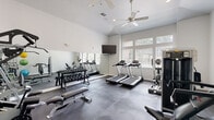 Located at 24011 Richards Road in Spring, TX, this single-story fitness center is designed to support a comprehensive workout experience. The space features vaulted ceilings that lend an open, airy feel, complemented by ceiling fans that promote air circulation throughout. Neutral gray walls and durable interlocking rubber flooring create a clean, functional aesthetic well-suited to an active environment.

Natural light flows generously into the space through a bank of large windows fitted with horizontal blinds, offering views of surrounding greenery while keeping the interior bright. A wall-mounted flat-screen television provides entertainment during cardio sessions.

The cardio area includes multiple treadmills and an elliptical trainer positioned near the windows. A full rack of dumbbells alongside a weight bench serves the free-weight zone, while a large wall mirror enhances spatial perception and allows users to monitor their form. Dedicated strength training stations, including a multi-function cable machine and additional benches, are positioned throughout. Medicine balls and exercise balls stored on a vertical rack round out the functional training options. The layout is thoughtfully organized to allow adequate space between equipment zones, supporting a safe and efficient workout environment.