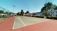 Located at 5280 Tamarack Cir E in Columbus, Ohio, this outdoor recreational facility centers around a full-size tennis court designed for active community use. The court features a hard surface playing area with clearly marked white boundary lines, complemented by a classic red surrounding border that defines the court's perimeter. Tall floodlights are positioned to support evening play, ensuring the space remains functional beyond daylight hours. The court is fully enclosed by chain-link fencing, providing both safety and structure to the playing environment. Lush mature trees and well-maintained hedgerows frame the space, offering natural shade and a pleasant green backdrop. The surrounding landscape opens to views of a neighboring residential community, integrating the court naturally into its setting. The single-level outdoor layout is easily accessible and straightforward in design, making it well-suited for recreational tennis at all skill levels. Under open skies and surrounded by established greenery, this facility offers a welcoming and functional outdoor space for residents and visitors to enjoy active recreation year-round.