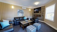 This single-floor space centers around a thoughtfully designed living area that balances comfort and contemporary style. The room features striped wallcovering in warm neutral tones, complemented by crisp crown molding that adds a polished architectural detail along the ceiling perimeter. A standout focal point is the accent wall finished in wood-look tile panels, which frames both a wall-mounted flat-screen television and a sleek, linear electric fireplace below — creating an inviting and visually striking entertainment hub. Seating is anchored by a dark leather sofa and matching armchair, accented with teal throw pillows that introduce a pop of color. A granite-topped coffee table sits at the center, adding a touch of elegance. Striped upholstered ottomans provide flexible additional seating. Wall sconces on either side of the room offer warm ambient lighting, while a flush-mount ceiling fixture provides general illumination. A ductless wall-mounted air conditioning unit ensures climate control. Abstract artwork adorns the walls, contributing to the overall refined yet relaxed ambiance of the space.