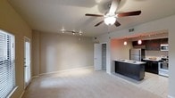 This single-floor space offers an open-concept layout that seamlessly blends the living area with the kitchen, creating a cohesive and functional environment. The living area is finished with plush carpeting and neutral-toned walls, lending a warm and inviting ambiance. A ceiling fan with an integrated light fixture serves as a central focal point while providing both air circulation and overhead lighting. Complementing this, a track lighting system along the ceiling offers additional directed illumination, making the space versatile for various lighting needs. The kitchen features a dark-toned breakfast bar that visually separates it from the living area without compromising the open feel. Dark cabinetry, a stainless steel refrigerator, and a pendant light above the bar contribute to a contemporary aesthetic. The kitchen flooring transitions to a hard surface, offering a practical contrast to the carpeted living space. A door leading to what appears to be a storage or utility closet is conveniently accessible from the main living area, adding to the functional design of the space. Overall, the layout and finishes reflect a clean, modern style suited for comfortable everyday living.
