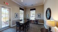 This single-floor community space functions as both a business center and a gathering area, thoughtfully designed to accommodate productivity and social interaction. The interior is finished with warm taupe walls accented by a contrasting chair rail, crisp white crown molding, and coordinating white baseboards, creating a polished and cohesive aesthetic throughout.

The space is divided into two functional zones. The primary gathering area centers on a substantial dark wood high-top table surrounded by black leather side chairs, ideal for meetings or collaborative work. A decorative round mirror with a dark wood frame adds visual interest to one wall, while framed artwork is distributed throughout, contributing to a refined atmosphere.

The business center zone features individual computer workstations — each equipped with a monitor and keyboard — positioned along the perimeter near large windows dressed with dark valances and horizontal blinds. These generously sized windows flood the space with natural light and offer views of the surrounding outdoor landscape.

Tall ceilings with recessed lighting enhance the sense of openness, while glass-paned interior French doors and a glass-paneled exterior door provide connectivity to adjacent spaces and the outdoors. The textured, tile-patterned flooring grounds the room with a durable and attractive finish suitable for high-traffic use.