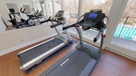 This fitness center is a bright, single-floor facility designed to support an active lifestyle. The space is outfitted with a selection of True-brand cardio equipment, including multiple treadmills, an elliptical trainer, and a recumbent stationary bike, providing a well-rounded range of aerobic workout options. Wood-style flooring runs throughout, offering a clean and durable surface underfoot, while a drop ceiling with recessed fluorescent lighting ensures consistent, even illumination across the room. A large wall mirror spans one side of the space, enhancing the sense of openness and allowing users to monitor their form during workouts. Expansive floor-to-ceiling windows wrap around a portion of the facility, flooding the interior with abundant natural light and offering direct views of an outdoor swimming pool and surrounding landscaped grounds. A glass door provides convenient access to the pool area. The warm, neutral-toned walls contribute to a welcoming atmosphere, and interior windows connect the fitness center to an adjacent laundry facility, adding a layer of practical convenience. Informational postings and workout charts are displayed along the walls, reinforcing the functional, community-oriented character of the space.