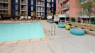 Located in the heart of Downtown Los Angeles, this dynamic outdoor amenity space at 555 N Spring St offers a well-appointed retreat within a colorful, modern residential complex. The centerpiece is a generously sized swimming pool with crystal-clear turquoise water, framed by a wide concrete pool deck outfitted with lounge seating, teal ottomans, yellow accent stools, and shaded cabana-style daybed structures. Lush succulent and agave plantings line the pool perimeter, adding a desert-modern aesthetic that complements the bold, multi-colored building facades surrounding the courtyard.

Adjacent to the pool area, an elevated rooftop terrace provides sweeping views of the Downtown Los Angeles skyline. This level features a mix of seating arrangements, including a hanging egg chair, wicker loungers, built-in sofa planters with succulents, and a dining table set — all set against a backdrop of the city's distinctive skyline. A stretch of artificial turf adds a soft, park-like quality to the rooftop setting.

Throughout the outdoor spaces, the design blends contemporary urban style with relaxed, resort-inspired comfort. Decorative planters, gravel landscaping, and accent lighting contribute to a cohesive and inviting atmosphere ideal for both social gatherings and quiet relaxation in the heart of the city.