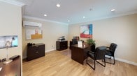 This single-story leasing office is designed to provide a professional and welcoming environment for staff and visitors alike. The entrance features a white paneled door with a fanlight transom window, flanked by two large windows dressed with horizontal blinds, allowing natural light to fill the reception area. Light wood-style flooring runs throughout, complementing the warm neutral tones of the walls and crisp crown molding that frames each room.

The main workspace is equipped with multiple dark wood desks, each accommodating a computer workstation, phone, and organizational accessories. Black leather executive chairs provide comfortable seating for staff, while guest chairs are positioned across the desks to facilitate resident or prospective tenant consultations. A vibrant abstract canvas artwork adds a decorative focal point to the primary office area.

Additional functional elements include a dedicated filing station with dark lateral file cabinets, a multifunction printer, and a corkboard for staff communications. Recessed ceiling lighting and a wall-mounted mini-split air conditioning unit ensure a comfortable working environment year-round. The overall layout is efficient and practical, reflecting a space well-suited for daily leasing operations.