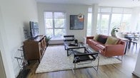 This single-floor space showcases a thoughtfully curated modern aesthetic, where clean lines and natural light come together to create an inviting atmosphere. The living area is a standout feature, anchored by rich hardwood flooring that runs throughout and a generous arrangement of floor-to-ceiling windows on multiple walls. These expansive windows flood the room with natural light while offering verdant outdoor views of the surrounding tree canopy, creating a seamless connection between the interior and the outdoors. The living space is well-appointed with a warm cognac leather sofa, sleek chrome-framed Wassily-style chairs, and a glass-top coffee table resting atop a plush cream shag rug — all contributing to a sophisticated yet approachable design. A sculptural arc floor lamp and curated wall art add personality and depth to the space. A wooden media console with open shelving provides both functional storage and a decorative anchor along one wall. The overall ambiance is one of airy, contemporary living — bright, open, and refined, with a careful balance of modern furniture and natural elements that make the space feel both stylish and livable.