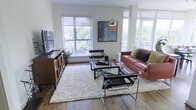 This single-floor space showcases a thoughtfully curated modern aesthetic, where clean lines and natural light come together to create an inviting atmosphere. The living area is a standout feature, anchored by rich hardwood flooring that runs throughout and a generous arrangement of floor-to-ceiling windows on multiple walls. These expansive windows flood the room with natural light while offering verdant outdoor views of the surrounding tree canopy, creating a seamless connection between the interior and the outdoors. The living space is well-appointed with a warm cognac leather sofa, sleek chrome-framed Wassily-style chairs, and a glass-top coffee table resting atop a plush cream shag rug — all contributing to a sophisticated yet approachable design. A sculptural arc floor lamp and curated wall art add personality and depth to the space. A wooden media console with open shelving provides both functional storage and a decorative anchor along one wall. The overall ambiance is one of airy, contemporary living — bright, open, and refined, with a careful balance of modern furniture and natural elements that make the space feel both stylish and livable.
