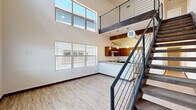 Located at 1571 North Pine Cliff Drive in Flagstaff, Arizona, this modern two-story home offers a thoughtfully designed layout across two levels. The main floor makes an immediate impression with soaring double-height ceilings and an abundance of natural light pouring in through large windows and clerestory glass. Light wood-look flooring runs throughout, complemented by dark wood baseboards that add a clean contrast to the crisp white walls. A ceiling fan anchors the bright, open living area. The kitchen is a standout feature, boasting rich wood cabinetry, granite-style countertops, a peninsula with a sink, stainless steel appliances, and stylish pendant lighting suspended from an exposed wood beam — all framed by the striking steel-railed staircase leading to the upper level. The upper floor houses two bedrooms, each featuring vaulted ceilings, sliding closet doors, and windows with neighborhood views. Three bathrooms serve the home, including an ADA-accessible bath with an open vanity, grab bars, and a roll-in shower, as well as a full bath with a tub-shower combo and granite-top vanity. The upper landing overlooks the living area below, reinforcing the home's open and airy character throughout.