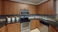 This single-floor residence showcases a cohesive design aesthetic defined by warm wood tones, dark granite surfaces, and neutral wall colors throughout. The kitchen is a functional and stylish centerpiece, featuring flat-front cabinetry in a rich wood finish, dark speckled granite countertops, and a gray tile backsplash. Stainless steel appliances, including an electric range with an over-the-range microwave and a dishwasher, provide a polished look. A double stainless steel sink sits beneath pendant lighting, and a breakfast bar connects the kitchen to the living and dining area, which features hardwood-style flooring and open, airy proportions.

The bedrooms are carpeted and finished with soft neutral tones, creating a calm and restful atmosphere. The primary bedroom accommodates a queen or king bed and includes a tufted upholstered headboard, mirrored nightstands, and warm ambient lighting. A secondary bedroom offers additional space with carpeting and room for seating. The bathroom mirrors the kitchen's design language, with a wood vanity cabinet, dark granite countertop, an undermount sink, and a wide wall-mounted mirror with a modern multi-bulb light fixture. A tub and shower combination completes the space.