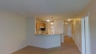 This single-floor residential space is thoughtfully laid out to maximize functionality and flow. The open-concept design connects the kitchen and living area seamlessly, creating a cohesive and practical living environment. The kitchen is equipped with warm wood-toned cabinetry, a built-in microwave over the range, a white electric stove, and ample counter space. A peninsula-style breakfast bar separates the kitchen from the adjacent living area, making it ideal for casual dining or entertaining. A chandelier-style light fixture adds a subtle decorative touch above the dining area, complementing the flush-mount ceiling lights throughout. Neutral-toned carpeting extends through the living areas and hallway, lending a soft, comfortable feel underfoot. The hallway, visible from the main living space, leads to the private areas of the home, with multiple doors suggesting well-defined, separate rooms. The consistent, light-colored walls throughout create a bright and clean atmosphere. Overall, the space offers a practical and comfortable single-level layout with a warm, understated aesthetic suited for everyday living.