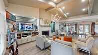 The Retreat at Brightside Clubhouse in Baton Rouge, Louisiana is a thoughtfully designed single-story community gathering space that blends comfort and modern style. The main lounge area serves as the heart of the clubhouse, boasting soaring vaulted ceilings with exposed wood beam detailing and a striking sputnik chandelier that anchors the room. A full-height textured fireplace creates a dramatic focal point, flanked by a large flat-screen TV mounted above teal horizontal shiplap shelving. Plush neutral seating, including sofas and armchairs accented with vibrant orange ottomans, provides an inviting atmosphere for residents to relax and socialize. Natural light filters throughout via large windows dressed with bold red-accented drapery. The kitchen area features granite countertops, a spacious island, gray shaker cabinetry, stainless steel appliances, and a mosaic tile backsplash. A rectangular pendant light fixture adds a contemporary touch above the island. French doors open to outdoor views, further brightening the space. A dedicated seating nook and leasing area complete the layout. Light wood-style flooring flows seamlessly throughout, unifying the space with a clean, modern aesthetic. Parcel lockers are also conveniently located on-site.