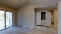This single-floor residence offers a clean and functional layout designed for comfortable everyday living. The main living area immediately draws attention with its vaulted ceiling, which adds a sense of openness and airiness to the space. Soft, neutral-toned carpet flooring runs throughout, providing a warm and inviting underfoot feel that complements the room's understated, neutral color palette on the walls. A ceiling fan fixture is installed overhead, offering both lighting and climate comfort. Natural light filters into the space through a window, brightening the interior with a calm, diffused glow. One of the standout practical features of this residence is the in-unit laundry area, thoughtfully integrated into a dedicated closet alcove. The bi-fold closet doors open to reveal a side-by-side washer and dryer, keeping laundry needs contained and accessible without sacrificing living space. An adjacent door leads further into the residence, suggesting additional rooms beyond the main living area. The overall ambiance of the space is calm and neutral, presenting a versatile canvas well-suited to a variety of furnishing styles and personal tastes. Clean lines, practical storage, and thoughtful amenities make this a highly functional and livable single-floor home.