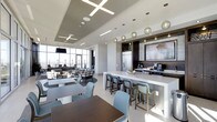 This contemporary single-floor space showcases a thoughtfully designed open-plan layout that seamlessly integrates a kitchen, dining, and lounge area into one cohesive environment. The architectural design features elevated ceilings with distinctive recessed tray detailing and cross-shaped linear LED lighting fixtures, lending a polished, modern aesthetic throughout.

The kitchen area is anchored by a sleek white waterfall island with bar-height seating in muted blue-grey upholstered chairs, complemented by dark cabinetry, a stainless steel refrigerator, and warm pendant globe lights that add an elegant contrast. A large wall-mounted display screen serves as a functional focal point within the kitchen zone.

Multiple dining tables with mixed chair configurations provide flexible seating arrangements, while a dedicated lounge area near the expansive floor-to-ceiling windows offers an inviting setting with ample natural light and panoramic views of the surrounding skyline. The light-toned flooring and neutral color palette—accented with dark wood tones—create a refined, professional ambiance throughout the space, making it well-suited for both casual gatherings and collaborative use.