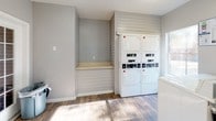 This single-floor laundry facility is designed to provide a clean, functional, and comfortable experience for its users. The space features warm hardwood-style flooring throughout, which pairs neatly with the neutral gray walls and bright white trim to create a tidy, welcoming atmosphere. A ceiling fan with a light fixture helps maintain air circulation within the room.

The facility is equipped with multiple coin-operated or card-operated washing machines and stacked dryer units, offering ample capacity for residents. Instructional signage is posted prominently above the machines, guiding users through the washing and drying process with ease. A built-in folding counter with a shiplap-style surround provides a convenient surface for sorting and folding laundry, accompanied by additional open shelf space.

Natural light flows generously into the space through several windows and a set of French doors that open to an outdoor area, keeping the environment bright and airy. The French doors also provide easy access and ventilation. Additional storage is available via a standard interior door and a closet, offering practical utility. The layout is open and spacious, allowing multiple users to move comfortably throughout the facility. Overall, the space strikes a balance between practical function and a clean, well-maintained aesthetic.
