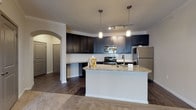 This single-floor residence offers a thoughtfully designed open-concept layout that seamlessly connects the kitchen and living areas. The kitchen is a standout feature, showcasing rich dark cabinetry that extends to the ceiling, granite countertops, a white subway tile backsplash, and a full suite of stainless steel appliances including a range, microwave, and refrigerator. A central island with a granite surface and undermount sink adds valuable prep space and serves as a natural dividing point between the kitchen and the living area. Pendant lighting and track lighting illuminate the kitchen, while a large pendant fixture brightens the adjacent living space. The living area features plush neutral carpeting and crown molding, with a ceiling fan adding both function and visual interest. An elegant arched passageway connects the main living space to the bedroom corridor. The bedrooms are generously sized, each outfitted with soft carpeting, ceiling fans, and windows with blinds that invite natural light. The bathroom features granite countertops, bold navy cabinetry, and wood-look tile flooring. Additional highlights include a dedicated laundry closet with washer/dryer hookups and ample storage throughout. The overall design balances modern finishes with a warm, comfortable ambiance.