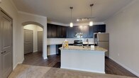 This single-floor residence offers a thoughtfully designed open-concept layout that seamlessly connects the kitchen and living areas. The kitchen is a standout feature, showcasing rich dark cabinetry that extends to the ceiling, granite countertops, a white subway tile backsplash, and a full suite of stainless steel appliances including a range, microwave, and refrigerator. A central island with a granite surface and undermount sink adds valuable prep space and serves as a natural dividing point between the kitchen and the living area. Pendant lighting and track lighting illuminate the kitchen, while a large pendant fixture brightens the adjacent living space. The living area features plush neutral carpeting and crown molding, with a ceiling fan adding both function and visual interest. An elegant arched passageway connects the main living space to the bedroom corridor. The bedrooms are generously sized, each outfitted with soft carpeting, ceiling fans, and windows with blinds that invite natural light. The bathroom features granite countertops, bold navy cabinetry, and wood-look tile flooring. Additional highlights include a dedicated laundry closet with washer/dryer hookups and ample storage throughout. The overall design balances modern finishes with a warm, comfortable ambiance.