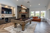 Located on 80th Street in Overland Park, Kansas, this single-story clubhouse offers a thoughtfully designed gathering space with a cohesive modern aesthetic. The entire space is unified by striking herringbone-patterned hardwood flooring that flows seamlessly from room to room, complemented by crisp white walls and vaulted ceilings that enhance the sense of openness.

The game and lounge area serves as a central highlight, anchored by a substantial billiard table positioned beneath a sculptural wood-and-metal pendant light. A floor-to-ceiling stacked stone fireplace with a warm wood mantel adds a dramatic focal point, while a wall-mounted flat-screen TV and a wet bar with dark cabinetry and a beverage cooler complete the entertainment setup. A leather sofa and large windows with outdoor views create a comfortable, relaxed atmosphere.

Adjacent to the game room, the main lounge features mid-century modern seating, decorative ring pendant lights, and an intricate laser-cut metal room divider that adds artistic dimension. A dedicated dining area includes a long communal bar-height table with leather counter stools, pendant lighting, and a decorative geometric accent wall in muted teal. A cozy booth nook with upholstered seating and a wall-mounted TV offers an intimate breakout spot. Natural light floods the space throughout.