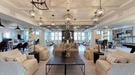 This single-story clubhouse offers a generously proportioned open gathering space defined by sophisticated yet comfortable design. The interior features a striking shiplap ceiling with exposed beams, anchored by a series of wrought-iron lantern-style pendant lights that cast warm, ambient illumination throughout. Large windows line the perimeter walls, flooding the space with natural light while framing views of the surrounding exterior. White brick accent walls add textural contrast and architectural character to the room. The main lounge area is thoughtfully arranged with plush, neutral-toned sofas and armchairs grouped around a substantial dark-finished wood coffee table, creating an inviting setting for relaxed conversation and socializing. Additional seating vignettes are positioned toward the rear of the space, offering quieter areas for smaller gatherings. A bar or dining area is visible in the background, extending the functionality of the clubhouse. The overall aesthetic blends farmhouse-inspired architectural elements with refined furnishings, resulting in a space that feels both welcoming and polished — well-suited for community events and everyday resident use.