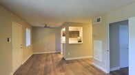 This single-floor residence offers a clean, functional layout designed for comfortable everyday living. The main living area is finished with warm hardwood-style flooring that extends throughout, creating a cohesive and inviting atmosphere. A ceiling fan overhead provides both comfort and a subtle design accent, while neutral-toned walls keep the space feeling bright and versatile.

A defining architectural feature is the pass-through opening that connects the living area to the kitchen, framed by a raised countertop that doubles as a breakfast bar. The kitchen is equipped with rich, dark wood cabinetry and track lighting mounted to the ceiling, lending it a warm and practical character. The textured ceiling adds a classic finish consistent throughout the space.

Additional practical details include a wall-mounted HVAC vent and a built-in wall unit, ensuring year-round climate control. The overall layout is open and unobstructed, allowing for flexible furniture arrangements and a sense of spaciousness. The interplay between the warm flooring tones and the neutral wall palette gives the space a grounded, welcoming ambiance suited to a variety of interior styles.