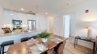 This single-floor space showcases a seamlessly integrated open-concept layout connecting the kitchen and dining area. The kitchen is outfitted with crisp white cabinetry, stone countertops with subtle veining, and stainless steel appliances including a French door refrigerator. A kitchen island with a undermount sink serves as both a functional workspace and a natural divider between the cooking and dining zones. Pendant lighting fixtures hang above the island, adding focused illumination and a contemporary design touch. Recessed lighting throughout ensures the space remains bright and well-lit. The dining area comfortably accommodates a full-sized wooden dining table surrounded by brown leather-style chairs, making it well-suited for everyday meals and entertaining. Light hardwood-style flooring runs throughout, contributing warmth and visual continuity. The neutral white walls and clean architectural lines create a modern, airy ambiance. Additional doors visible in the background suggest access to further rooms, adding to the functional versatility of this well-designed space.