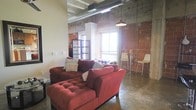 This single-floor space showcases a compelling industrial aesthetic that blends raw architectural elements with comfortable living. The living area is anchored by bold red tufted sofas that contrast beautifully against the exposed brick walls, which serve as a defining feature throughout the space. Polished concrete floors add to the industrial character while remaining practical and visually striking.

The ceilings soar overhead, revealing exposed ductwork and metal conduit piping that emphasize the loft-style design. A pendant barn light hangs from above, complementing the utilitarian aesthetic. Natural light streams in through large windows, brightening the space and highlighting the textural contrast between the smooth painted walls and the raw brick.

A small dining or bar area near the windows includes slim bar-height chairs, making efficient use of the open floor plan. The combination of warm brick tones, deep red furnishings, and industrial fixtures creates an inviting yet edgy ambiance throughout, offering a well-balanced mix of character and comfort in a thoughtfully designed single-level layout.