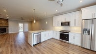 Located at 121 Starlight Walk in Holbrook, NY, this single-story home showcases a thoughtfully designed open-concept layout finished with consistent warm-toned wood flooring and crown molding throughout. The kitchen serves as a central hub, featuring white shaker cabinetry, light stone countertops, a subway tile backsplash, and a peninsula with an undermount sink and pendant lighting overhead. Stainless steel appliances—including a gas range, built-in microwave, dishwasher, and French door refrigerator—complete the well-appointed space, which flows seamlessly into the adjoining living and dining areas. The living room is anchored by a striking floor-to-ceiling stacked-stone fireplace, adding texture and warmth to the neutral palette. Ceiling fans are present throughout, enhancing comfort. The bedrooms offer generous proportions, natural light from multiple windows, and include walk-in closets with built-in shelving. Bathrooms are elegantly finished with marble-look tile, white shaker vanities, stone countertops, and glass-enclosed tub-shower combinations. The primary bathroom features a dual-sink vanity and a spacious walk-in tiled shower. The overall aesthetic is clean, contemporary, and move-in ready.