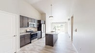 Located in Marinette, Wisconsin, this thoughtfully designed single-floor residence offers a clean, modern aesthetic with quality finishes throughout. The open-concept layout is anchored by a well-appointed kitchen featuring dark shaker-style cabinetry, granite-look countertops, stainless steel appliances including a range, microwave, and refrigerator, and a central island with a built-in sink and pendant lighting overhead. Soaring vaulted ceilings amplify the sense of openness, while large sliding glass doors at the rear flood the living area with natural light and provide outdoor access. Light hardwood-style flooring runs seamlessly throughout the entire space, lending a cohesive, contemporary feel. The bedroom is a comfortable, well-proportioned retreat featuring vaulted ceilings, a ceiling fan, a window with views of the surrounding greenery, and baseboard heating. A dedicated closet offers convenient storage. The bathroom is clean and functional, showcasing a walk-in shower with a white surround, a dark wood vanity with stone-look countertop, and matching light flooring. A hallway connects the private areas of the home, completing this efficient and stylish single-level layout.