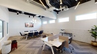 This thoughtfully designed single-floor office space seamlessly blends industrial and contemporary aesthetics to create a productive and visually appealing work environment. The space is defined by its striking exposed black ceiling with visible ductwork and linear LED lighting, which contrasts beautifully against the crisp white walls and whitewashed brick wainscoting that runs throughout.

The open coworking area features modular white workstations with privacy dividers and ergonomic chairs, offering flexible seating for multiple occupants. A herringbone-patterned carpet tile floor adds a refined, textural element underfoot. High clerestory windows line the upper walls, drawing in natural light while maintaining privacy, and the generous ceiling height contributes to an airy, open atmosphere.

A dedicated conference area accommodates a large rectangular table surrounded by navy blue upholstered chairs with gold accents, anchored by a striking globe chandelier overhead. A vibrant tropical-themed artwork adorns the accent wall, adding a warm focal point to the meeting space. Interior glass partitions allow visual connectivity between zones while preserving a sense of structure. The space is fully equipped with office essentials including a printer station, making it a functional and well-appointed professional environment.