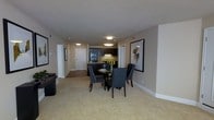 This single-floor residence offers an open and functional layout that seamlessly connects the dining area and kitchen. The dining space is anchored by a round dark-wood table surrounded by four upholstered chairs with light trim detailing, creating an elegant yet approachable setting. A framed abstract artwork on the adjacent wall adds a decorative focal point to the area.

The kitchen, visible from the dining space, features dark espresso cabinetry paired with under-cabinet lighting that illuminates the countertops. Black appliances complement the rich cabinetry tones, contributing to a cohesive, contemporary aesthetic. A hallway extends toward what appears to be additional rooms, including a door that likely leads to a bedroom or utility space, with a second framed piece of wall art adding character along the corridor.

Soft, light-toned carpeting covers the main living and dining areas, adding warmth underfoot and contrasting gently with the darker furnishings. The walls are finished in a warm neutral beige, and flush-mount ceiling lights provide consistent, even illumination throughout. The textured ceiling and clean white baseboards complete the polished, well-maintained interior feel of this space.