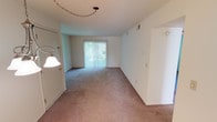 This single-floor residence offers a straightforward and functional layout designed for comfortable everyday living. The entry opens directly into a generously sized living room, finished with wall-to-wall carpet in a neutral tone and illuminated by a ceiling-mounted chain light fixture. A sliding glass door fitted with vertical blinds fills the space with natural light and provides access to the outdoors. The living area flows toward a dining space, highlighted by an elegant hanging chandelier that adds a decorative touch. The kitchen is efficiently arranged in an L-shaped configuration, featuring white cabinetry, a double-basin stainless steel sink positioned beneath a window, a coil-burner electric range, and a full-size refrigerator, all set against light-toned countertops and vinyl flooring. The bedrooms are carpeted and bright, with at least one room benefiting from a window that admits natural light and outdoor views. The bathroom includes a full tub and shower combination enclosed by sliding glass doors with a chrome frame, complemented by clean, neutral-toned walls and tile. Throughout the unit, the consistent use of light, warm wall colors and plush carpeting creates a cohesive and welcoming ambiance suited for a range of living arrangements.