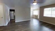 This single-floor space features an open-concept layout that seamlessly connects the main living area, dining space, and kitchen. Dark hardwood-style flooring runs throughout, providing a rich contrast against the neutral, light-toned walls and crisp white baseboards.

The living area is outfitted with a ceiling fan and light fixture, offering both comfort and illumination. It flows naturally into a bright dining area, which is highlighted by a decorative chandelier and a large window fitted with white blinds that allows natural light to fill the room. A wall-mounted HVAC unit serves the dining area, providing climate control convenience.

The kitchen, partially visible from the main living space, features dark cabinetry and a stainless steel refrigerator, giving it a modern, functional feel. The open sightlines between rooms contribute to a sense of spaciousness throughout the unit.

Overall, the space presents a clean, updated aesthetic with practical finishes, making it a functional and comfortable single-level living environment.