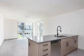 This contemporary single-floor unit showcases a thoughtfully designed open-concept layout with clean lines and a bright, neutral palette throughout. The kitchen is a standout feature, boasting a large central island with a striking stone countertop and an integrated undermount sink paired with a matte black industrial-style faucet. Light wood cabinetry lines the walls alongside a full suite of stainless steel appliances, including a French door refrigerator, range, and built-in microwave, all complemented by a herringbone tile backsplash. Recessed lighting illuminates the space efficiently. The living area is generously sized, filled with natural light streaming through expansive floor-to-ceiling sliding glass doors that open to views of the surrounding neighborhood and lush greenery. Light-toned wood-look flooring runs throughout, adding warmth and continuity. The bedroom continues the airy aesthetic with its own large sliding glass door, a ceiling fan, and a baseboard heater, ensuring year-round comfort. The bathroom features a clean, all-white tub-shower surround with subway-style tiling, maintaining the unit's modern and minimalist character. A hallway connects the spaces seamlessly, with glimpses of outdoor views visible even from the interior corridor.