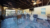 Located at 3919 Carnegie Ave in Myrtle Beach, South Carolina, this single-level covered outdoor pavilion is designed for communal gathering and outdoor entertainment. The structure features an exposed dark wood beam ceiling with a vaulted roofline, lending the space a rustic yet polished character. Ceiling fans are mounted throughout, ensuring comfortable airflow during warm coastal days.

The pavilion's flooring consists of multi-toned slate-style tiles that add texture and visual interest underfoot. A prominent stone-clad bar serves as a central feature, equipped with bar-height seating and a wall-mounted flat-screen television above, making it ideal for social gatherings and watching events. Multiple high-top tables and chairs are arranged throughout the space, comfortably accommodating groups of various sizes.

Large open-air wall openings on multiple sides of the pavilion provide unobstructed views of the surrounding pool area and lush landscaping, while seamlessly connecting the interior to the outdoors. Built-in gas grills positioned near one of the open sides offer convenient outdoor cooking capabilities directly accessible from the seating area.

Coastal-themed artwork on the walls and abundant natural light flowing in from the open sides enhance the relaxed, beach-inspired ambiance of this well-appointed community gathering space.