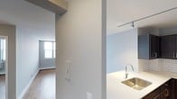 This single-floor space showcases a clean and contemporary design characterized by a neutral color palette of crisp whites and soft grays throughout. The open-concept layout connects the kitchen and living areas seamlessly, creating a sense of flow and spaciousness. The kitchen is equipped with a modern stainless steel sink set into a light-toned countertop, complemented by a sleek, arc-style faucet. Overhead, a track lighting system provides targeted, adjustable illumination, adding a practical yet stylish touch to the space. The living area benefits from natural light streaming in through a large window, brightening the hardwood-style flooring and giving the room an airy, welcoming atmosphere. Structural columns are integrated into the layout, adding architectural character while subtly defining different zones within the open floor plan. The walls are finished in smooth, light tones that enhance the sense of brightness and openness. Overall, the space presents a minimalist and functional design, well-suited for those who appreciate modern interiors with thoughtful architectural details and a cohesive, understated aesthetic.