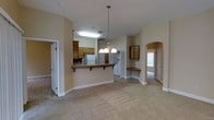 This single-floor home offers a thoughtfully designed open-concept layout that seamlessly connects the main living areas. The kitchen is a functional centerpiece, featuring warm honey-toned wood cabinetry, a dark stone-finish countertop, and a raised peninsula with a convenient breakfast bar overhang — ideal for casual dining or entertaining. A white refrigerator and recessed fluorescent lighting keep the kitchen bright and practical. Overhead, a classic multi-bulb chandelier hangs gracefully above the peninsula, adding a touch of elegance to the space. The open living area extends from the kitchen and is finished with plush, neutral-toned carpeting that lends a warm and comfortable feel throughout. A dedicated laundry area is conveniently tucked nearby, adding to the home's practical layout. One of the standout architectural features is the use of graceful arched doorways and passageways, which add a refined, classic character to the interior. A built-in nook with additional cabinetry and a bench seat provides extra storage and a cozy corner detail. Multiple doorways lead to additional rooms, suggesting a well-connected and functional floor plan. The warm beige walls and consistent neutral palette create a cohesive, inviting ambiance throughout the home.