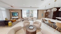Located at 15350 SW 136th St in Miami, FL, this sophisticated residence lounge is designed to offer a refined communal living experience on a single expansive floor. The open-concept layout seamlessly connects multiple distinct seating and gathering zones, all unified by large-format white tile flooring and a bright, neutral color palette accented with warm wood tones and gold-finish details.

The lounge area features plush, curved cream-toned armchairs arranged around a hexagonal wood coffee table, alongside a more relaxed living zone with a deep charcoal sofa, textured area rug, and a wall-mounted flat-screen TV set against a patterned accent wall. An artistic copper-toned chandelier adds a striking focal point overhead. Sheer white floor-to-ceiling curtains frame oversized windows that flood the space with natural light and offer views of the surrounding greenery and building exterior.

A built-in banquette seating area with rustic wood tables provides an additional casual gathering spot directly beside the windows. The kitchen and bar area is a standout feature, boasting rich dark wood cabinetry, a long white quartz island with bar seating, globe pendant lighting in brushed brass, stainless steel appliances, and open shelving. A full-length mirror wall enhances the sense of depth and space, while glass entry doors connect the lounge to the outdoors.