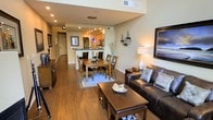 This thoughtfully designed 2-floor residence offers a seamless open-concept layout that connects the living, dining, and kitchen areas into one cohesive and inviting space. Rich hardwood flooring flows throughout, lending warmth and continuity to the interior.

The living area is anchored by a comfortable leather sofa, complemented by classic wooden coffee and side tables, and tasteful wall art that adds a coastal-inspired character to the space. Recessed lighting and a stylish table lamp create a layered, welcoming ambiance.

The dining area sits adjacent to the living room, centered around a generous wooden dining table with seating for six, positioned beneath a decorative area rug. Pendant lighting adds a refined touch overhead.

The kitchen features warm wood cabinetry, stainless steel appliances, and a peninsula with bar seating — ideal for casual dining or entertaining. Striking blue pendant lights serve as a bold focal point, adding a modern contrast to the warm tones throughout.

Neutral, earthy wall tones and consistent finishes across both levels give the space a cohesive and polished aesthetic. The open sightlines from the main living area into the kitchen and beyond create a sense of spaciousness, making the residence feel airy and functional for everyday living.