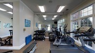 This fitness center is thoughtfully designed to accommodate a range of workout needs within a single-floor layout. The space features crown molding and white-trimmed walls, giving it a polished, finished appearance that sets it apart from a typical gym environment. Recessed rectangular fluorescent lighting fixtures illuminate the room evenly, complementing the natural light that streams in through multiple windows along one wall, offering views of surrounding greenery.

A full-length mirror spans one wall, visually expanding the space and allowing users to monitor their form during exercise. The flooring consists of a patterned carpet, providing a comfortable and cushioned surface underfoot. Cardio equipment, including elliptical trainers and additional machines, is arranged along the windowed side of the room, while a multi-tiered dumbbell rack stocked with a comprehensive range of free weights is positioned along the opposite wall. A flat weight bench and a blue exercise mat offer additional options for strength and flexibility training. A wall-mounted television is also present, adding convenience for users during workouts. A glass-paneled entry door provides easy access and contributes to the room's open, airy feel.