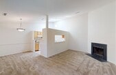This single-floor residence offers a thoughtfully arranged layout with a clean, neutral aesthetic throughout. The open living and dining area is generously proportioned, featuring warm neutral carpet underfoot and a large window dressed with vertical blinds that invites natural light and views of surrounding greenery. A half-wall partition with a pass-through window visually connects this space to the kitchen, enhancing the open feel while maintaining defined zones.

The galley-style kitchen is well-appointed with light wood cabinetry lining both walls, offering ample storage and counter space. White appliances include a range and refrigerator, complemented by light hardwood-style flooring. Notably, the kitchen also accommodates an in-unit washer and dryer, adding a high degree of everyday convenience. Soaring ceilings with globe pendant lighting give this space an airy, open character.

The residence includes two carpeted bedrooms, each finished with neutral tones and windows providing natural light and outdoor views. The full bathroom features a tiled shower-tub combination, hardwood-style flooring, and a wood-trimmed vanity. A separate half bathroom offers a pedestal sink and mirror, providing additional functionality for guests.