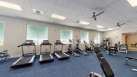 This fitness center offers a comprehensive workout environment designed to support a wide range of exercise routines. The space is laid out across a single open floor, finished with durable blue patterned carpet throughout and bright white walls that contribute to a clean, welcoming atmosphere. The drop ceiling features recessed panel lighting and multiple ceiling fans, ensuring the space remains well-lit and ventilated during workouts.

Large windows along the perimeter walls flood the interior with natural light while providing views of the surrounding exterior, creating an open and airy feel. The cardio zone is well-stocked with multiple treadmills, elliptical trainers, stationary bikes, and a rowing machine, offering plenty of options for aerobic training.

The strength training area includes a variety of Nautilus and TKO cable and selectorized machines, a multi-functional power rack, a punching bag, and a dumbbell rack with an adjustable bench. Full-length mirrors line one wall to assist with form and technique. A dedicated functional fitness corner features stability balls, medicine balls, resistance bands, and exercise mats stored on a Smart Fitness rack. The facility also includes a separate adjoining room visible through a doorway, adding additional utility to this fully appointed fitness space.