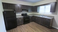 This single-floor space is designed with practicality and comfort in mind, offering a cohesive layout that makes efficient use of its footprint. The kitchen serves as a standout feature, showcasing rich dark espresso-toned cabinetry that extends from floor to ceiling, providing generous storage throughout. Light-toned countertops offer a pleasant contrast against the dark cabinetry, creating a balanced and visually appealing workspace. The kitchen is fully equipped with black appliances, including a gas range with oven, an overhead range hood, and a full-sized refrigerator. A double-basin stainless steel sink is positioned beneath a window, allowing natural light to fill the space during the day. The warm wood-look plank flooring runs throughout, adding a sense of warmth and continuity to the interior. The walls are finished in a neutral light tone, complementing the overall palette and keeping the space feeling bright and open. The kitchen layout is configured in an L-shape, maximizing counter space and promoting ease of movement. Together, these elements create a functional and welcoming environment well-suited for everyday living.