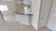 This single-floor residence offers a clean, functional layout designed for comfortable everyday living. The open-concept design connects the living area seamlessly to the kitchen, creating a bright and cohesive space. A peninsula-style breakfast bar with a dark granite-look countertop serves as the natural dividing point between the two areas, offering both additional prep space and casual seating potential. The living area features soft, neutral-toned carpeting that adds warmth underfoot, while the kitchen transitions to tile flooring for a practical finish. The kitchen is equipped with white cabinetry and a full-size refrigerator, complemented by overhead fluorescent lighting that keeps the space well-lit. A charming porthole-style window in the kitchen area adds a distinctive architectural touch, introducing natural light into the space. The walls are finished in a light, warm beige throughout, contributing to an airy and inviting atmosphere. A ceiling fan in the living area provides added comfort, and a thermostat on the wall reflects a climate-controlled environment. The entryway is marked by a classic white paneled door with brushed hardware, completing the tidy and well-maintained character of this single-level home.
